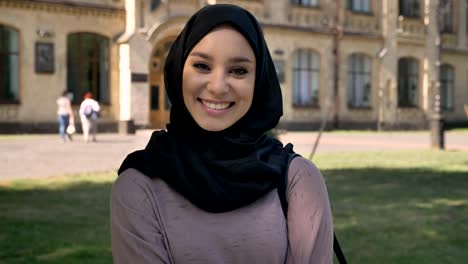 Young-sweet-muslim-girl-in-hijab-is-standing-and-smiling-in-daytime-in-summer,-watching-at-camera,-building-on-background,-religiuos-concept,-emotional-concept