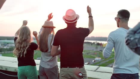 Rear-view-group-of-people-enjoying-beautiful-city-landscape-raising-arms-on-rooftop-party-at-sunset