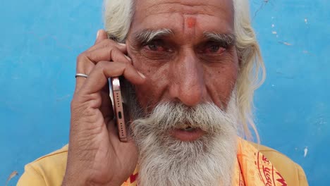 Hindu-Sadhu-Heiliger-Mann-mit-grauem-Bart-sitzt-und-spricht-ruhig-auf-seinem-Handy