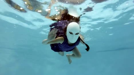 A-child-in-a-white-fairy-mask-swims-and-poses-underwater-in-a-purple-dress,-looks-at-the-camera-and-waving-his-hands-on-a-blue-background.-Slow-motion.