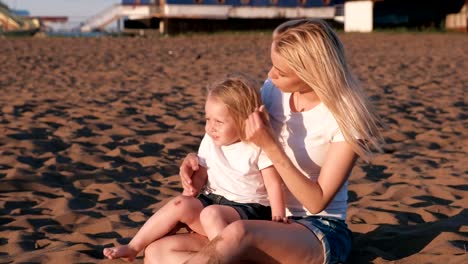 Beautiful-blonde-mom-and-daughter-play-with-sand-sitting-together-on-the-beach.