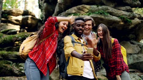 Glücklicher-afroamerikanischer-Kerl-macht-Videoanruf-mit-Smartphone-während-der-Wanderung-im-Wald-mit-Freunden,-junge-Menschen-sind-auf-der-Suche-am-Bildschirm,-winken-Hand-und-sprechen.