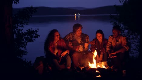 Happy-and-relaxed-guys-and-girls-are-singing-while-their-friend-is-playing-the-guitar-near-campfire-in-forest-near-lake-at-night.-Nature,-recreation-and-music-concept.