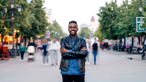 Zoom-out-time-lapse-of-emotional-African-American-man-standing-in-street-with-arms-crossed-and-looking-at-camera-when-men-and-women-are-whizzing-by.