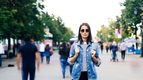 Time-lapse-portrait-of-confident-young-lady-in-sunglasses-standing-in-city-center-among-running-people-and-resting-looking-at-camera.-Time-and-youth-concept.