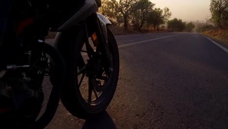 cinematic-shot-of-a-motorcycle-rider-in-a-sunset-time-crossing-the-road-in-the-forest,-seeing-some-many-parts-of-the-motorcycle-and-the-sky-and-the-trees,-camera-is-taking-the-wheel-crossing-the-road-between-the-fog