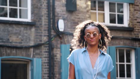 Moda-joven-mujer-negra-vestida-con-vestido-azul-y-gafas-de-sol-caminando-pasado-cámara-en-la-calle,-iluminado,-ángulo-bajo