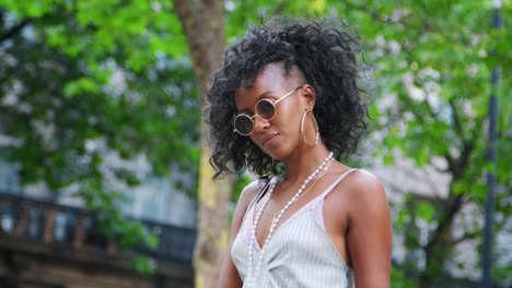 Moda-joven-mujer-negra-vestida-con-camiseta-y-gafas-de-sol-redondas-de-pie-en-la-calle,-árboles-en-el-fondo