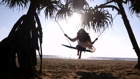 Young-happy-woman-in-swimsuit-and-shirt-relaxing-at-swing-at-tropical-ocean-beach.-Beautiful-girl-sitting-on-swing-and-enjoying-summer-vacation-or-holiday.-Female-have-fun-at-sea-shore.-Rear-back-view