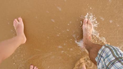 Point-of-view-of-young-couple-stepping-together-at-the-golden-sand-at-sea-beach.-Male-and-female-legs-walking-near-ocean.-Bare-feet-of-pair-going-on-sandy-shore-with-waves.-Summer-vacation-Closeup-POV