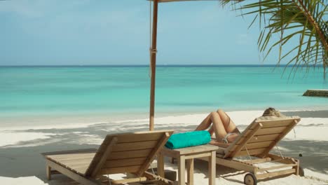 Close-up-of-Beautiful-Blonde-Female-Lying-on-the-Deck-Chair-on-the-Beach.-Sunbathing-on-the-Exotic-Location-with-Turquoise-Sea-in-the-Background.