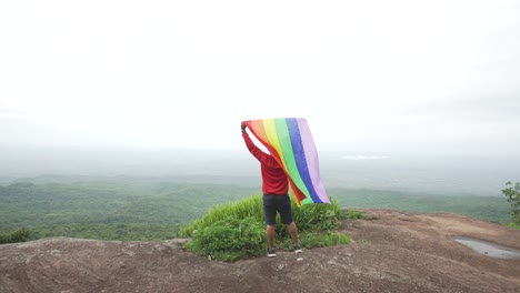 Mann-erhöhen-Regenbogen-Farbe-LGBTI-Fahnenschwingen-in-harten-Wind-auf-Mountain-Top-Sicht