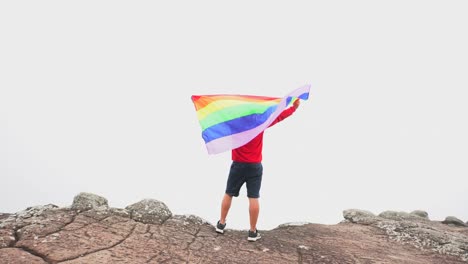 Mann-erhöhen-Regenbogen-Farbe-LGBTI-Fahnenschwingen-in-harten-Wind-auf-Mountain-Top-Sicht