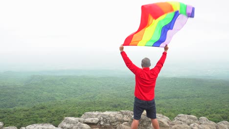 Mann-erhöhen-Regenbogen-Farbe-LGBTI-Fahnenschwingen-in-harten-Wind-auf-Mountain-Top-Sicht