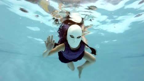 A-little-girl-in-a-fabulous-white-mask-swims-and-poses-under-the-water,-looks-at-the-camera-and-waving-his-hands.