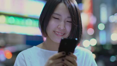 Portrait-of-the-Attractive-Japanese-Girl-and-Wearing-Casual-Clothes-Uses-Smartphone.-In-the-Background-Big-City-Advertising-Billboards-Lights-Glow-in-the-Night.