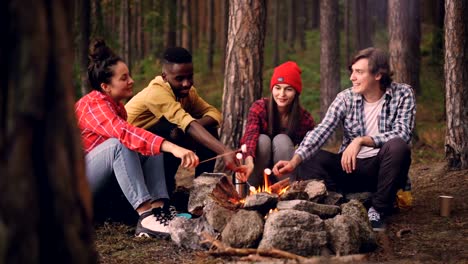 Hungry-travelers-are-cooking-marshmallow-on-fire-and-eating-it-from-sticks-during-conversation-around-campfire,-people-are-talking-and-laughing-enjoying-sweet-food.