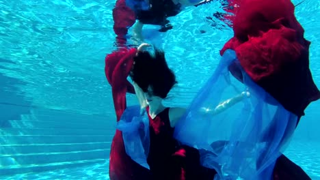 Beautiful-girl-bride-in-red-dress-swims-underwater-in-outdoor-pool-with-red-and-blue-cloth-in-her-hands.-She-looks-at-the-camera-and-smiles.