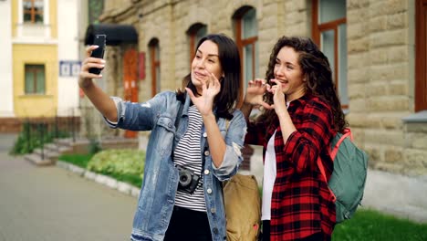 Cheerful-girls-foreign-travelers-are-taking-selfie-using-smartphone-standing-outdoors-and-posing-with-hand-gestures-showing-v-sign-and-heart-with-fingers-and-laughing.