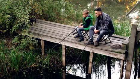 Good-looking-man-and-his-cute-son-are-fishing-in-pond-from-wooden-dock-sitting-on-chairs-with-rods-and-talking.-Loving-family,-common-hobby-and-generations-concept.