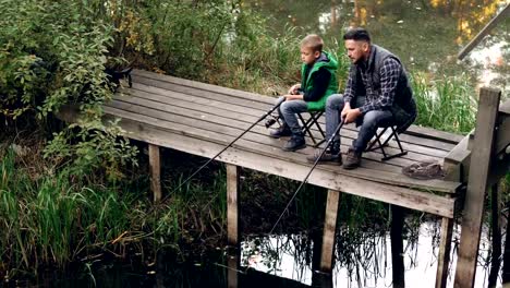 Gut-aussehender-Mann-und-Sohn-niedlich-sind-Fischen-im-Teich-aus-hölzernen-Dock-sitzen-auf-Stühlen-mit-Stangen-und-reden.-Liebevolle-Familie,-gemeinsames-Hobby-und-Generationen-Konzept.