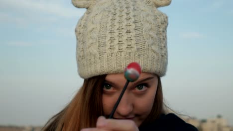 Pretty-teenage-girl-in-gray-knitted-hat-eating-lollipop-in-front-of-camera-and-smiling,-close-up-shot,-outdoor