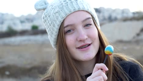 Hermosa-adolescente-en-una-gorra-de-punto-gris,-que-muestra-y-comiendo-piruleta,-artística-y-expresiva-tocando-en-la-cámara,-metraje-de-acciones.