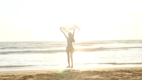 Young-happy-woman-with-scarf-running-on-the-ocean-beach-at-sunset.-Girl-enjoying-summer.-Female-in-bikini-with-flying-scarf-in-hands-jogging-on-the-sea-shore.-Close-up