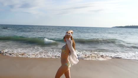 Mujer-feliz-en-bikini-y-camiseta-caminando-por-la-playa-cerca-del-océano-y-levantando-la-mano.-Joven-hermosa-chica-disfrutando-de-la-vida-y-la-diversión-a-la-orilla-del-mar.-Vacaciones-de-verano-o-vacaciones-de-paisaje-en-el-fondo