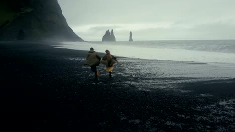 Back-aerial-view-of-young-hipster-couple-running-on-the-black-volcanic-beach-near-the-troll-toes-in-Iceland