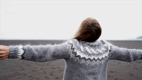 Junge-Frau-ausgestreckten-durch-das-Meer-stehend-am-schwarzen-Sandstrand,-Haare-im-Wind-Island---weiblich-verspielt-genießen-Natur-und-Freiheit---Slow-Motion-läuft