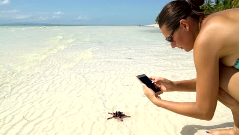 Una-mujer-en-un-traje-de-baño-toma-imágenes-de-una-estrella-de-mar-en-un-smartphone