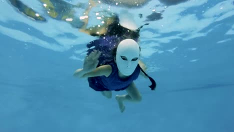 An-unusual-little-girl-swims-and-poses-underwater-in-a-fabulous-white-mask-and-purple-dress,-looks-at-the-camera-and-waving-her-hands-on-a-blue-background.-Slow-motion.