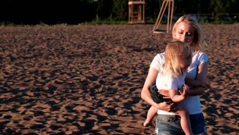 Schöne-blonde-Mutter-und-Tochter-kuscheln-und-sprechen-am-Strand-bei-Sonnenuntergang.