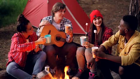 Fröhliche-Junge-Menschen-Freunde-sind-klirrende-Gläser-mit-Getränken-sitzen-um-Feuer-im-Wald-mit-warmen-Marshmallow-auf-Sticks,-Lächelnder-Mann-hält-Gitarre.