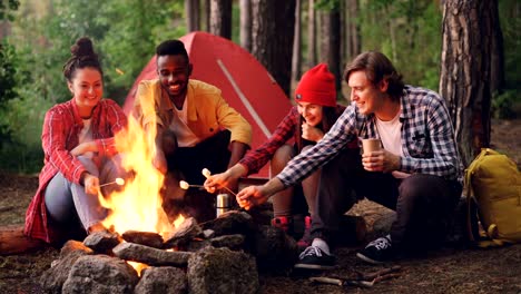 Happy-men-and-women-diverse-group-are-cooking-food-on-fire-at-campsite-sitting-around-campfire-and-holding-sticks-with-marshmallow.-Camping-and-friendship-concept.