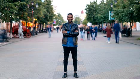 Time-lapse-retrato-de-alegre-hombre-del-afroamericano-de-pie-en-el-centro-con-ropa-elegante-mirando-a-cámara-y-sonriendo-mientras-las-personas-pasan.