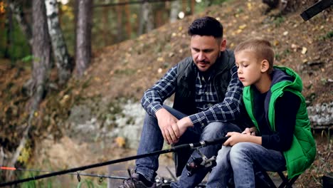 Handsome-guy-loving-dad-is-teaching-his-son-to-catch-fish-sitting-on-river-bank-together,-boy-is-holding-rod-and-moving-it-talking-to-daddy.-Hobby,-adventure-and-family-concept.