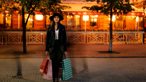 Zoom-out-time-lapse-of-serious-young-lady-in-fashionable-garments-standing-outdoors-in-pedestrian-street-holding-shopping-bags-and-looking-at-camera,-busy-people-are-passing-by.