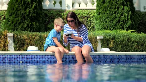 Young-mother-taking-photo-of-little-son-using-professional-camera-on-edge-swimming-pool