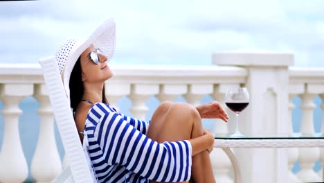 Elegant-woman-in-sunglasses-and-hat-at-balcony-sitting-on-chair-hugging-legs-by-crossed-hands