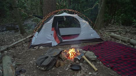 Bushcraft-camp-site-in-the-wilderness-with-campfire,-axe,-and-blanket.