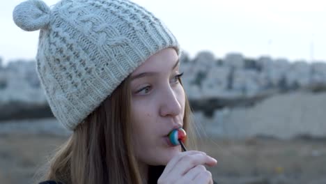 Beautiful-teenage-girl-in-a-gray-knitted-cap,-which-showing-and-eating-lollipop,-artistic-and-expressive-playing-on-camera,-outdoor-footage.