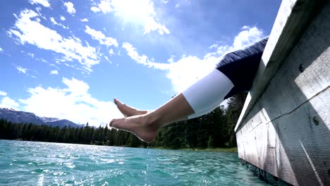 Woman's-feet-dangle-from-wooden-pier,-above-lake