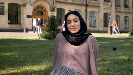 Young-happy-muslim-girl-in-hijab-is-sitting-on-lawn-in-sun,-watching-at-camera,-builging-on-background,-religious-concept,-relax-concept