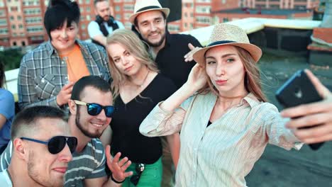 Elegant-young-woman-in-hat-taking-selfie-surrounded-by-friends-using-smartphone-at-rooftop-party