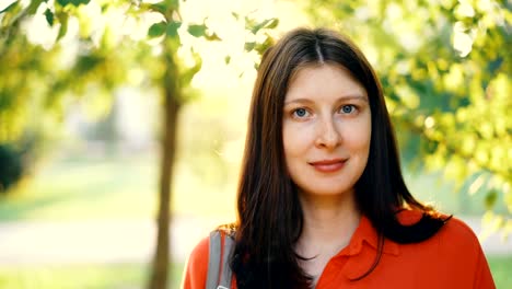 Close-up-portrait-of-attractive-young-woman-looking-at-camera-and-smiling-standing-in-park-on-sunny-summer-day.-Green-leaves,-grass-and-sunlight-are-visible.