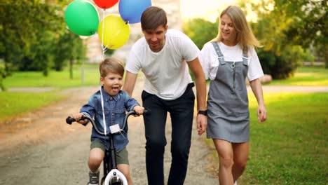 Loving-parents-are-teaching-son-cute-boy-to-ride-bike-in-city-park,-child-is-cycling-and-his-father-is-holding-him-helping-to-balance-while-mother-is-walking-beside-them.