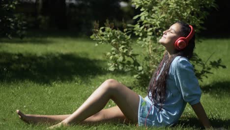 Junges-Mädchen-mit-langen-Haaren,-anhören-von-Musik-über-Kopfhörer-mit-Smartphone-sitzen-auf-dem-Rasen-im-Park-bei-sonnigem-Wetter.-4K
