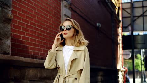 Attraktive-Frau-mit-blonden-Haaren-spricht-auf-dem-Handy-zu-Fuß-entlang-der-Straße-in-modernen-Stadt.-Junge-Dame-trägt-trendige-schwarze-Sonnenbrille-und-Sommerfell.